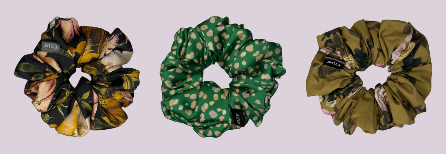 Three silk scrunchies from the brand Atila & Co are displayed side-by-side on a solid white background. The left scrunchie features a black, yellow, and brown floral pattern, the middle scrunchie is green with beige polka dots, and the right scrunchie is olive green with a subtle floral print.